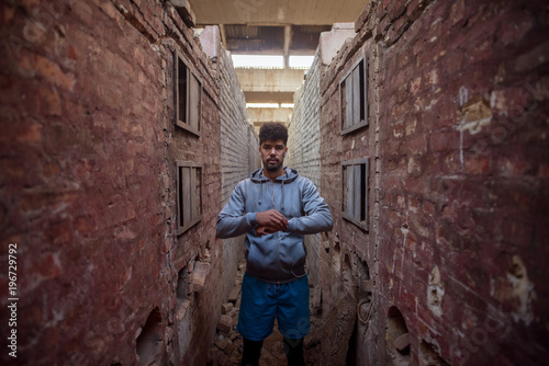 Fototapeta Naklejka Na Ścianę i Meble -  Portrait of focused motivated afro-american young handsome sportive man with earphones standing inside of the abandoned place in the middle of two walls and thinking.