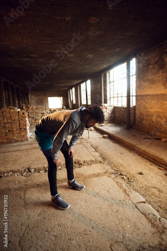Portrait of active afro-american young attractive athletic man with earphones crouching and resting after workout inside of the abandoned place.
