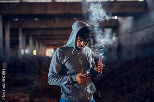 Portrait of joyful afro-american young handsome man relaxing inside of the abandoned hangar place with earphones and cigarette while choosing music on a mobile.