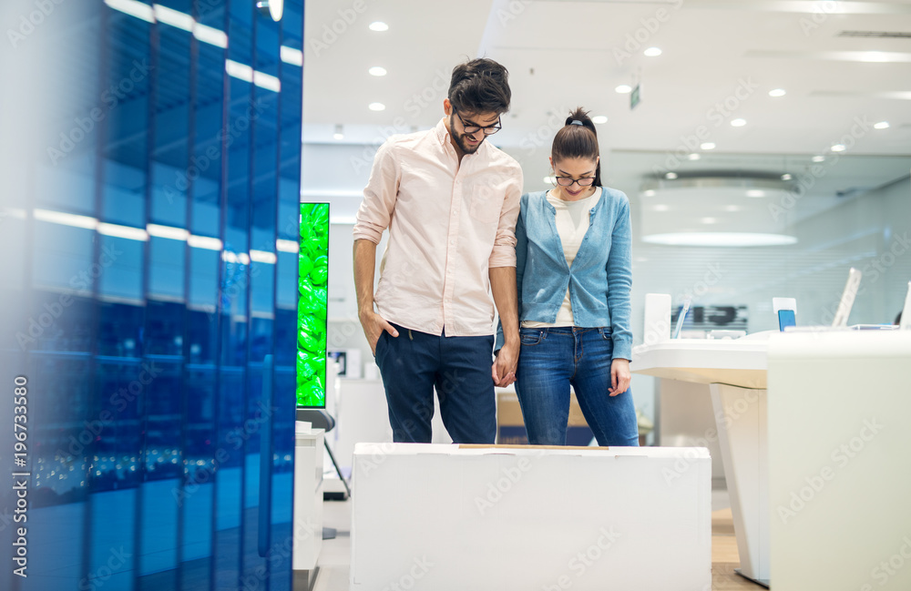Young happy love couple standing behind big cartoon box on the floor and looking down after buying a new electronic product in the tech store. Young happy love couple standing behind big cartoon box on the floor and looking down after buying a new electronic product in the tech store.