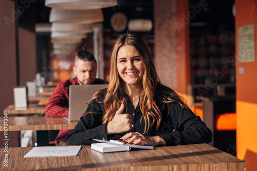 business woman sitting by the table in cafe with notebook while showing thumb up and looking at the camera