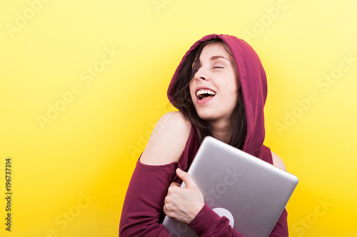 Smiling student with closed eyes holds a laptop in hands on yellow background in studio