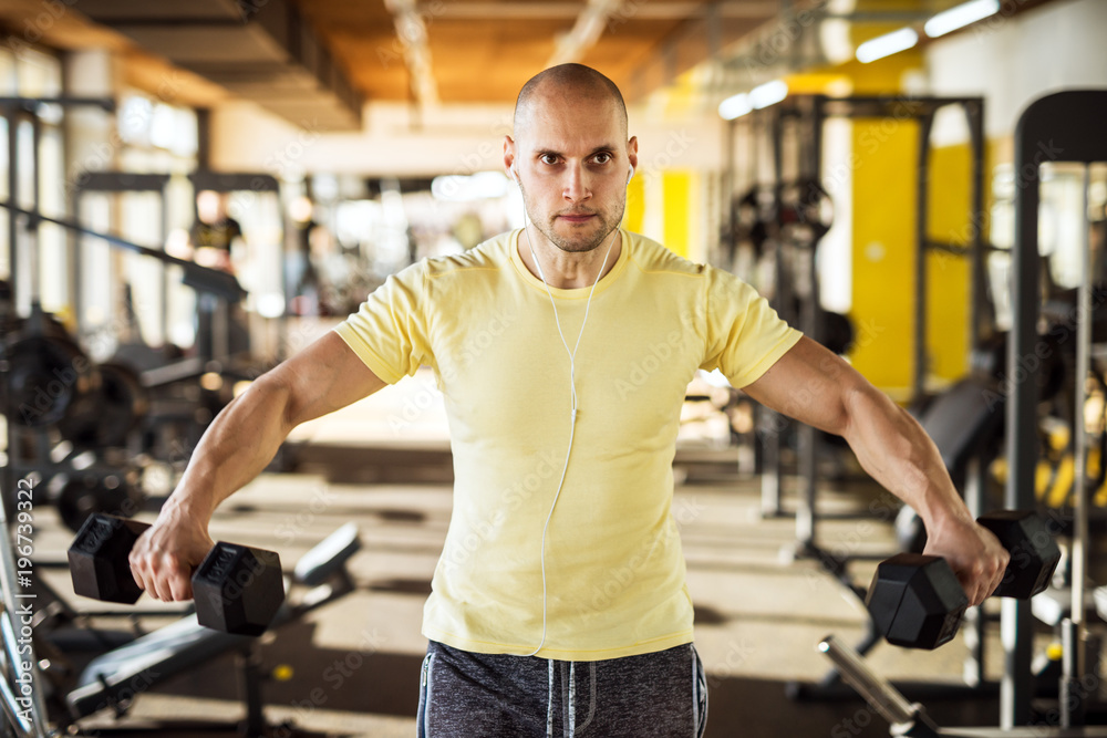 Portrait of strong muscular active healthy young man raising dumbbells with open arms and listening music in the gym. Portrait of strong muscular active healthy young man raising dumbbells with open arms and listening music in the gym.