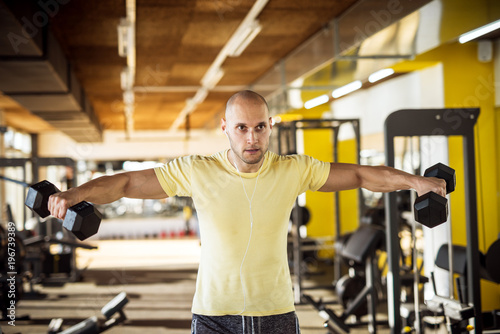 Portrait of strong muscular active healthy young man raising dumbbells with open arms and listening music in the gym.