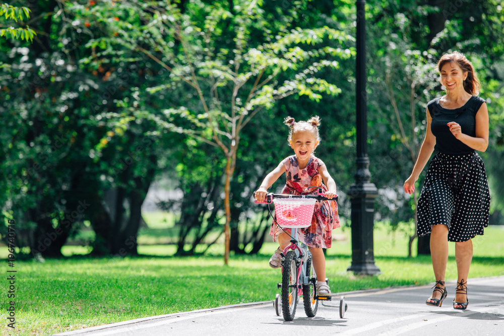 Beautiful and happy young mother teaching her cute daughter to ride a bicycle. Both smiling and looking at each other. Summer park in background. Happy parenting concept.