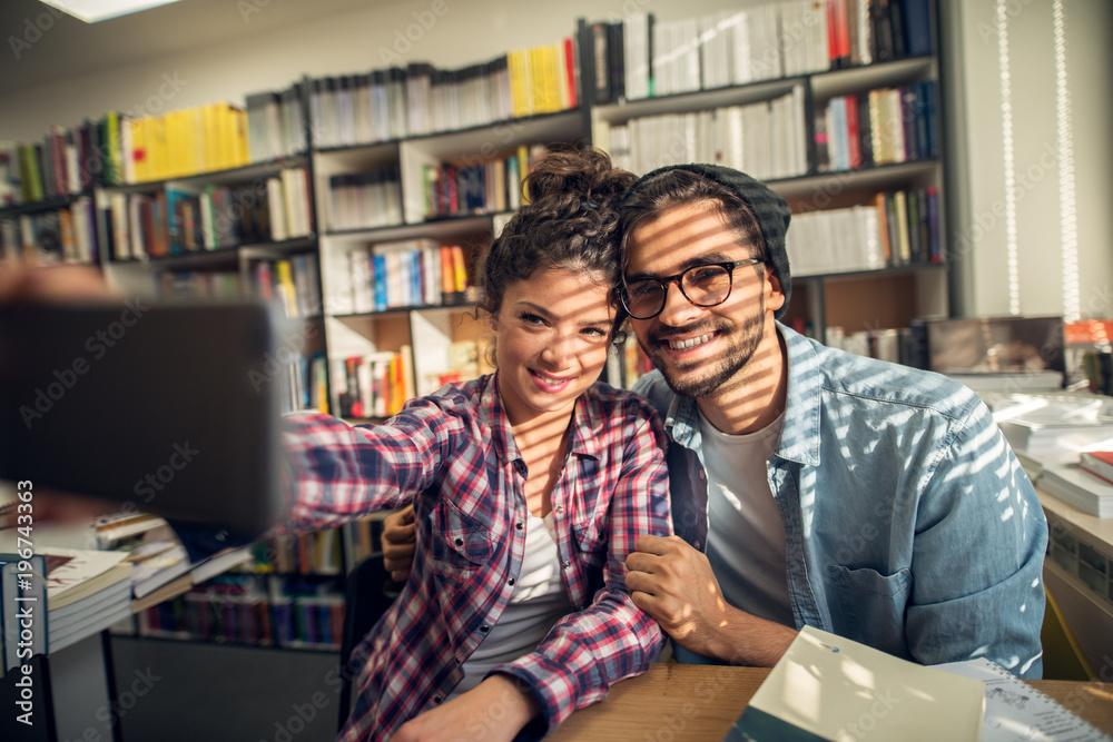 Close up of smiling hipster cheerful young student love couple sitting in the high school library and taking a selfie near the sunny window. Close up of smiling hipster cheerful young student love couple sitting in the high school library and taking a selfie near the sunny window.