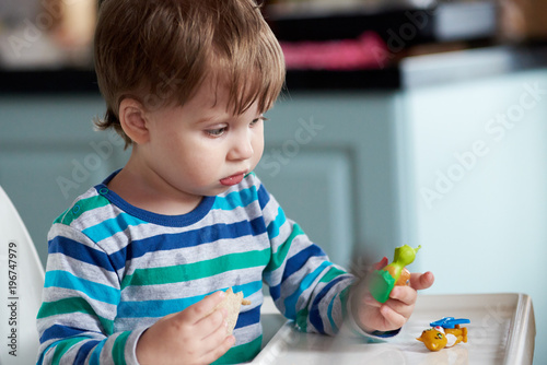 Cute european boy is sitting in the baby chair at home. He is playing with a toy and eating. biscuit.