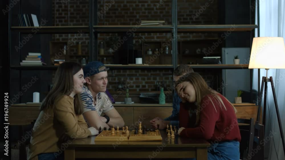Excited young woman going for checkmate in game of chess using white king to knock over opposing piece while playing chess with her female friend at home. Happy girl defeating her friend in chess game Excited young woman going for checkmate in game of chess using white king to knock over opposing piece while playing chess with her female friend at home. Happy girl defeating her friend in chess game