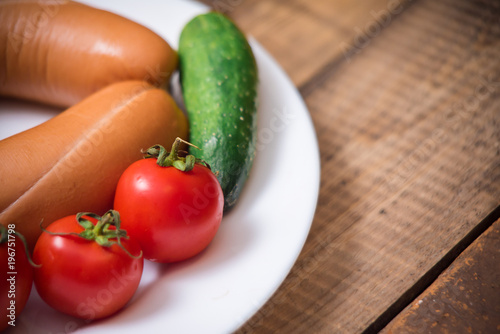 Cherry tomatoes, cucumbers and raw sausages on white plate on wooden table closeup