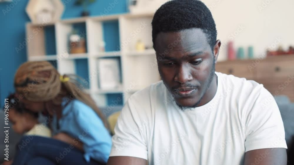 Close up view of young African American man feeling bad, rubbing his forehead, being sick. Woman playing with child on the background. Troubles, stress. Slow motion Close up view of young African American man feeling bad, rubbing his forehead, being sick. Woman playing with child on the background. Troubles, stress. Slow motion