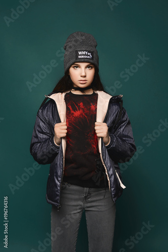 Young brunette teen model girl, in jacket and hat, stands over dark-green background, isolated