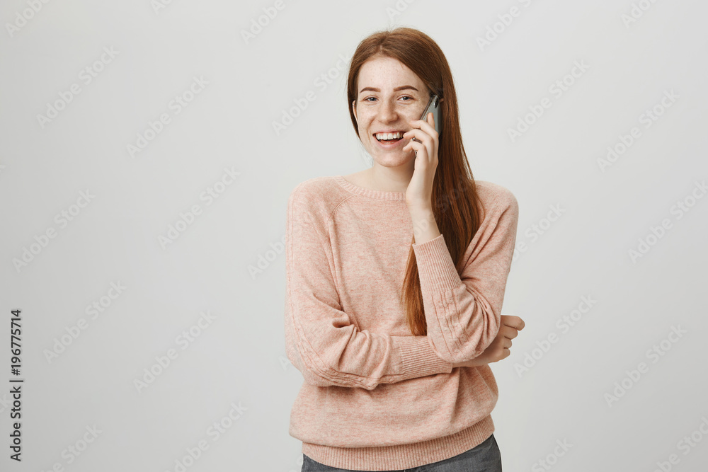 Positive interesting redhead girl talking on smartphone while laughing and looking with positive expression at camera, crossing chest with one hand, standing against gray background. Positive interesting redhead girl talking on smartphone while laughing and looking with positive expression at camera, crossing chest with one hand, standing against gray background.