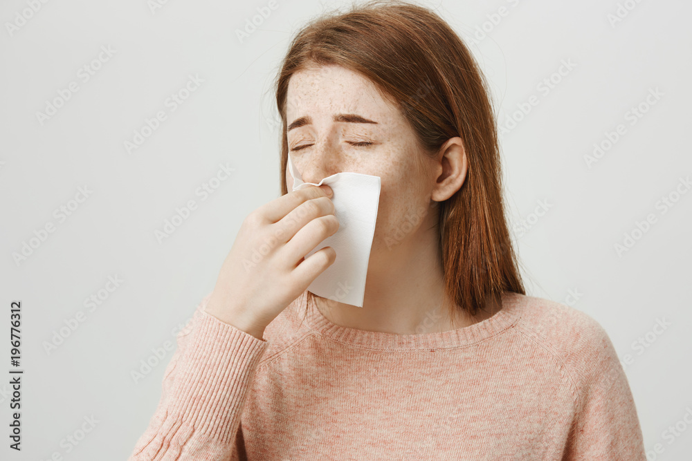 Close-up portrait of upset redhead girl having cold or flu, blowing nose in napkin while frowning and feeling bad, standing against gray background. Woman is bothered with allergy and runny nose Close-up portrait of upset redhead girl having cold or flu, blowing nose in napkin while frowning and feeling bad, standing against gray background. Woman is bothered with allergy and runny nose