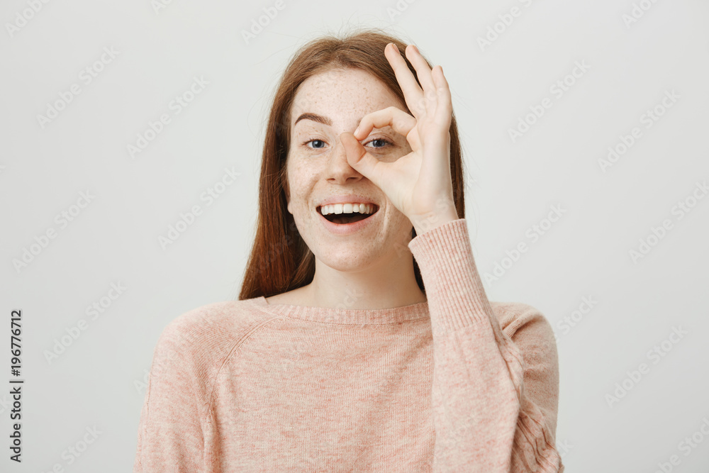 Studio portrait of positive cute redhead woman showing okay sign over eye and looking through it at camera while smiling cheerfully and standing against gray background. I am watching you Studio portrait of positive cute redhead woman showing okay sign over eye and looking through it at camera while smiling cheerfully and standing against gray background. I am watching you