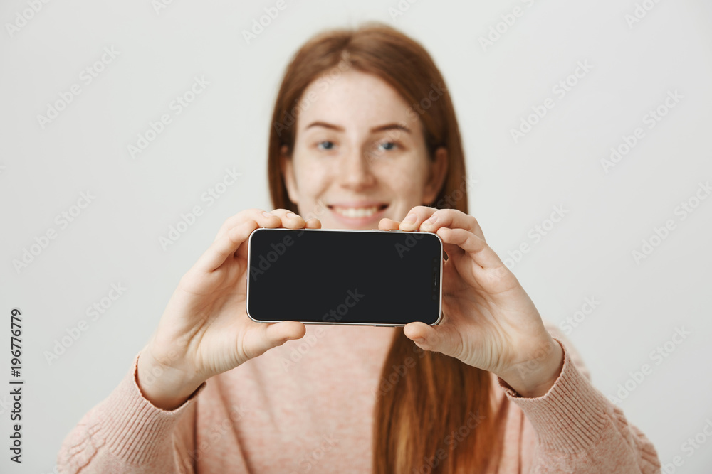 Blurred portrait of cute caucasian redhead model holding smartphone with both hands, advertising it over gray background. Seller shows new model of device to potential customer