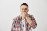 Waist-up portrait of joyful charming male in glasses and with trendy haircut, covering mouth, trying to hold laugh, standing against gray background. Guy put braces on teeth and shy to show it