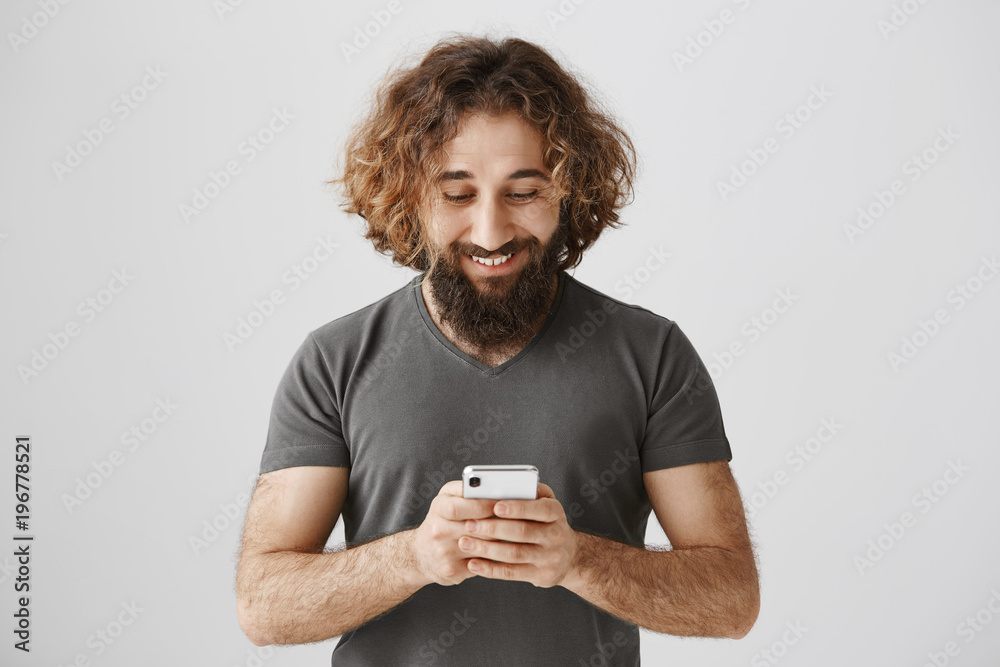 Best way of communication. Portrait of pleased and concentrated eastern male holding smartphone, playing games or messaging, grinning from excitement, reading positive news about his company Best way of communication. Portrait of pleased and concentrated eastern male holding smartphone, playing games or messaging, grinning from excitement, reading positive news about his company