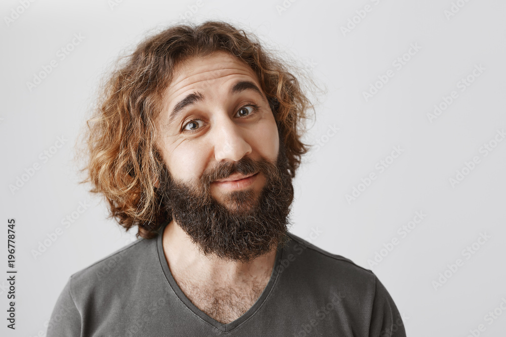 Indoor portrait of doubtful polite eastern guy with beard tilting and lifting eyebrows in disbelief, listening explanation of daughter, smiling with uncertain expression over gray background