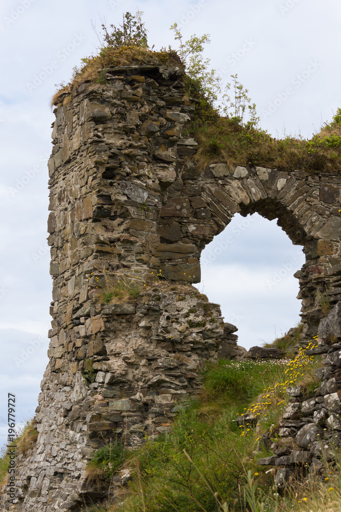 Stromeferry, Scotland - June 10, 2012: Closeup of Window in rock wall of Castle Strome ruins on green hill. Gray blue sky. Green weeds on wall. Yellow flowers up front.