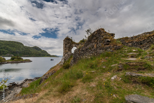 Stromeferry, Scotland - June 10, 2012: Part of Castle Strome ruins on green hill. Green weeds on walls. Gray Loch Carron. Forested hill on oposite shore. Dark rainy cloudscape.