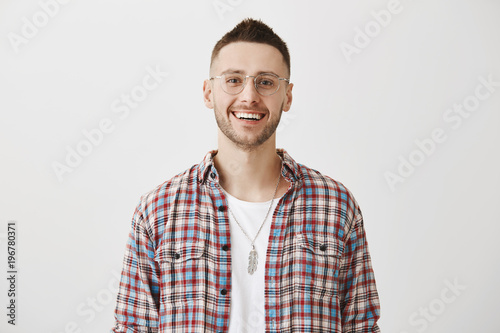 Always nice to talk with smart person. Indoor shot of attractive interesting caucasian guy smiling broadly while standing against gray background and discussing new inventions with friend