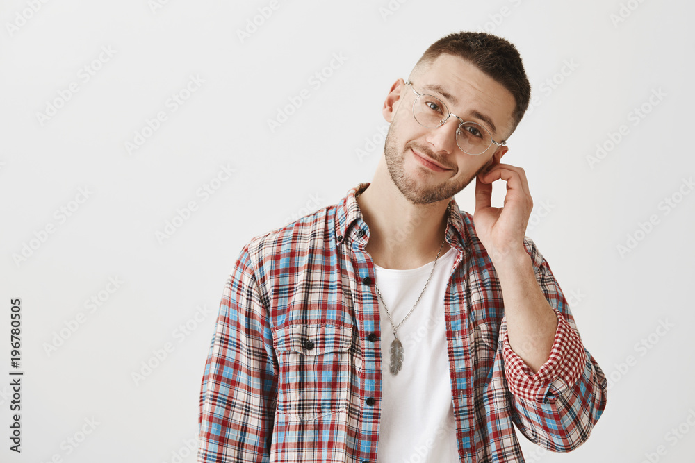 Indoor shot of pleasant good-looking man with beard in transparent glasses, rubbing ear and tilting head right, smiling and feeling tired while standing over gray background. Emotions concept Indoor shot of pleasant good-looking man with beard in transparent glasses, rubbing ear and tilting head right, smiling and feeling tired while standing over gray background. Emotions concept