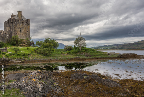 Dornie, Scotland - June 10, 2012: The waters of the lochs and Brown-stone entire Eilean Donan Castle. Under rain-heavy dark cloudscape. Low tide exposes brown seaweed. Wide shot shows hills on horizon