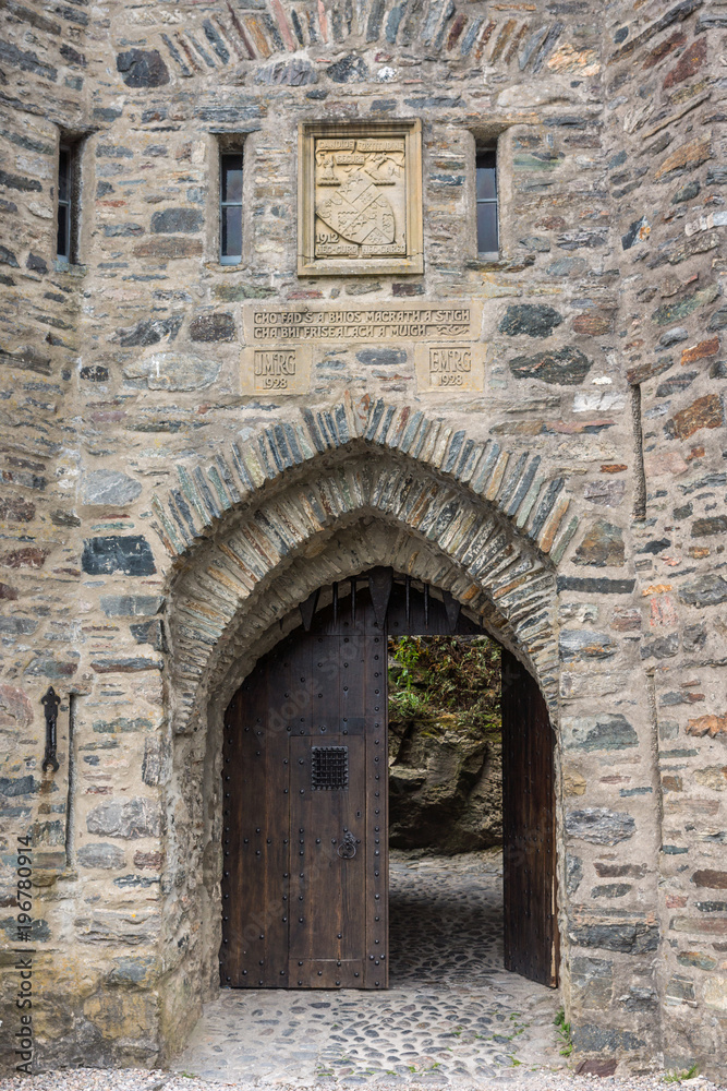 Dornie, Scotland - June 10, 2012: Closeup of gray-stone bowed entrance to Eilean Donan Castle with beige coat of arms above. Brown door open showing some green vegetation.
