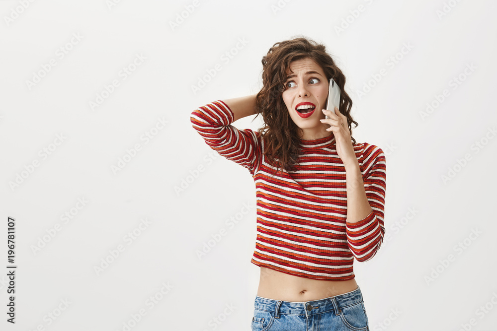 Girlfriend received call from guy while cheating on him. Troubled good-looking female in trendy striped cropped-top and red lipstick, scratching head and talking on smartphone, being confused