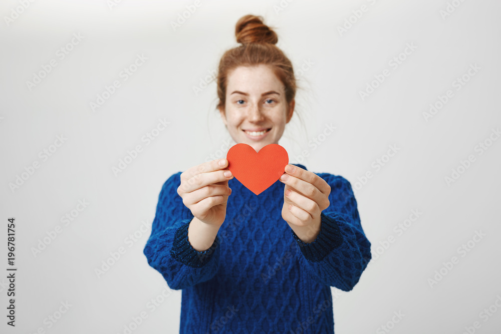 Keep it safe in your hands. Portrait of charming young woman in love pulling paper heart towards camera and smiling cheerfully while offering to start relationship to person she admires
