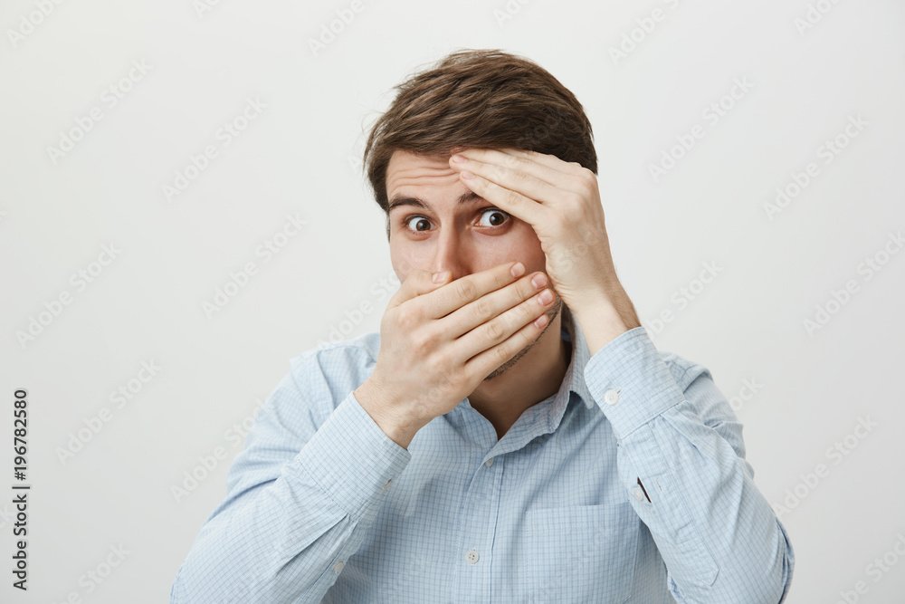 Is it over now. Portrait of scared and shocked young attractive man covering face with hands as if hiding or feeling fear, peeking at camera through hole, standing against gray background Is it over now. Portrait of scared and shocked young attractive man covering face with hands as if hiding or feeling fear, peeking at camera through hole, standing against gray background