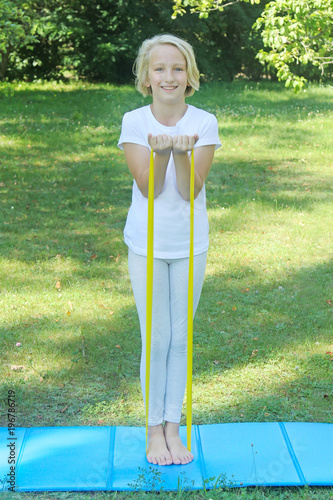 Beautiful school aged kid girl playing sports with a latex band and mat outdoors in the park 