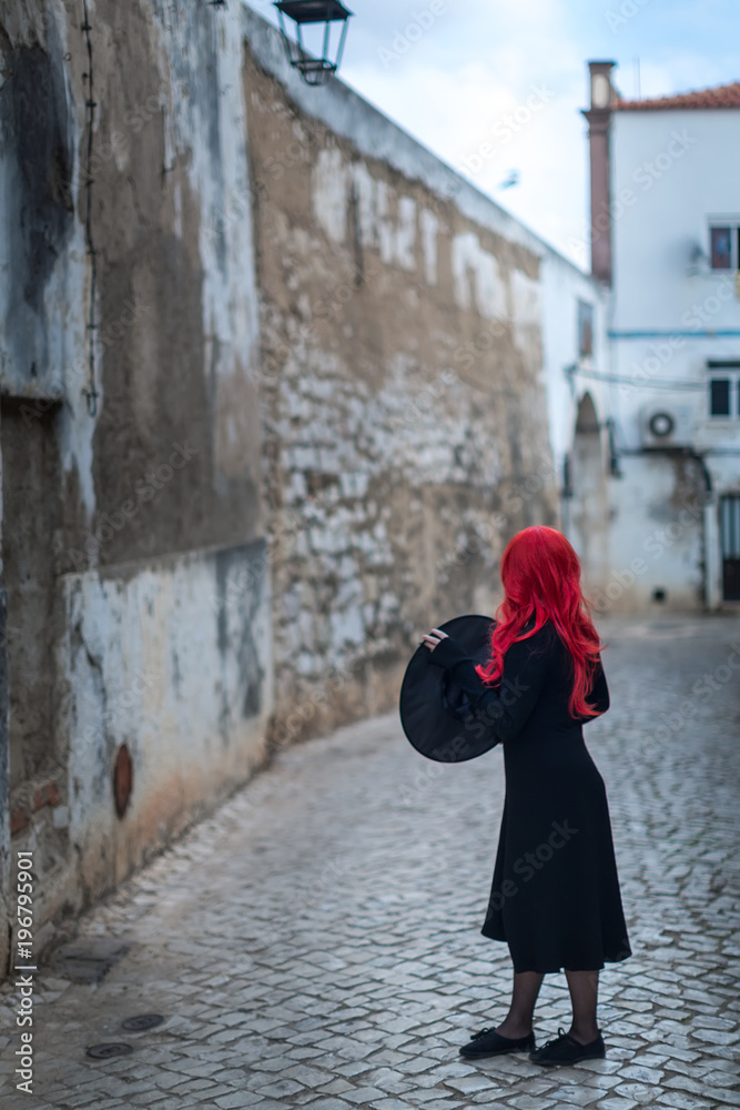 A small witch in a black dress with red hair is holding a hat in her hands and standing with his back against the backdrop of the street of the old city A small witch in a black dress with red hair is holding a hat in her hands and standing with his back against the backdrop of the street of the old city