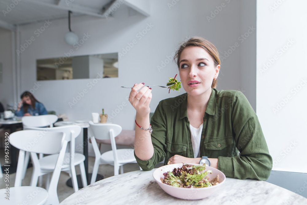 Beautiful young woman sitting in a cozy light restaurant with a fork in her hands, eating salad and looking away. Good food at the restaurant. Beautiful young woman sitting in a cozy light restaurant with a fork in her hands, eating salad and looking away. Good food at the restaurant.