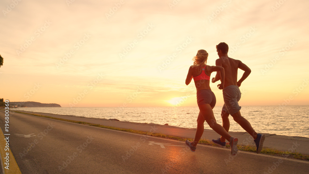COPY SPACE: Unrecognizable young couple jogs down ocean road at stunning sunset.