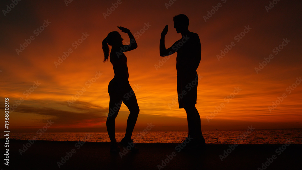 LOW ANGLE, SILHOUETTE: Happy young man and woman high five at stunning sunset.