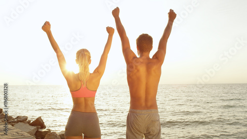 LENS FLARE: Unrecognizable fit couple look at sea and lift their arms overhead.
