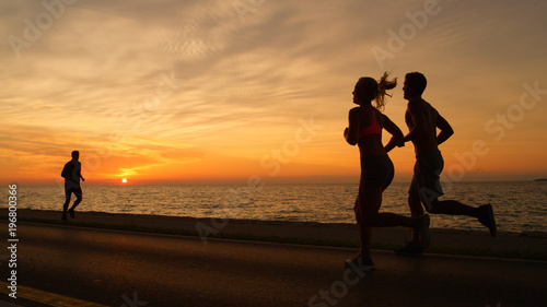 SILHOUETTE: Fit people run along silent ocean road in front of summer sunset.