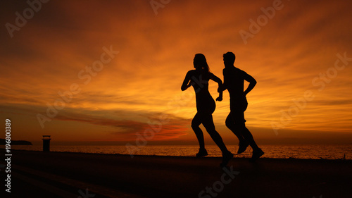 SILHOUETTE: Unknown athletic couple running along calm ocean at orange sunset.