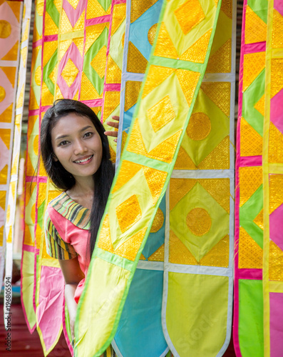 Beautiful woman peeks out of the wall from the colorful ornamentals curtains in the Buddhist monastery. A cheerful Asian girl is hiding behind decorative hangings.