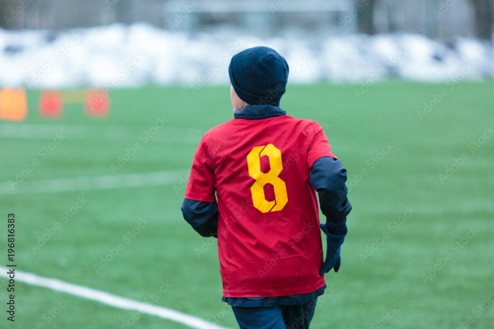  boy in red uniform -football player plays soccer on the green field in the winter