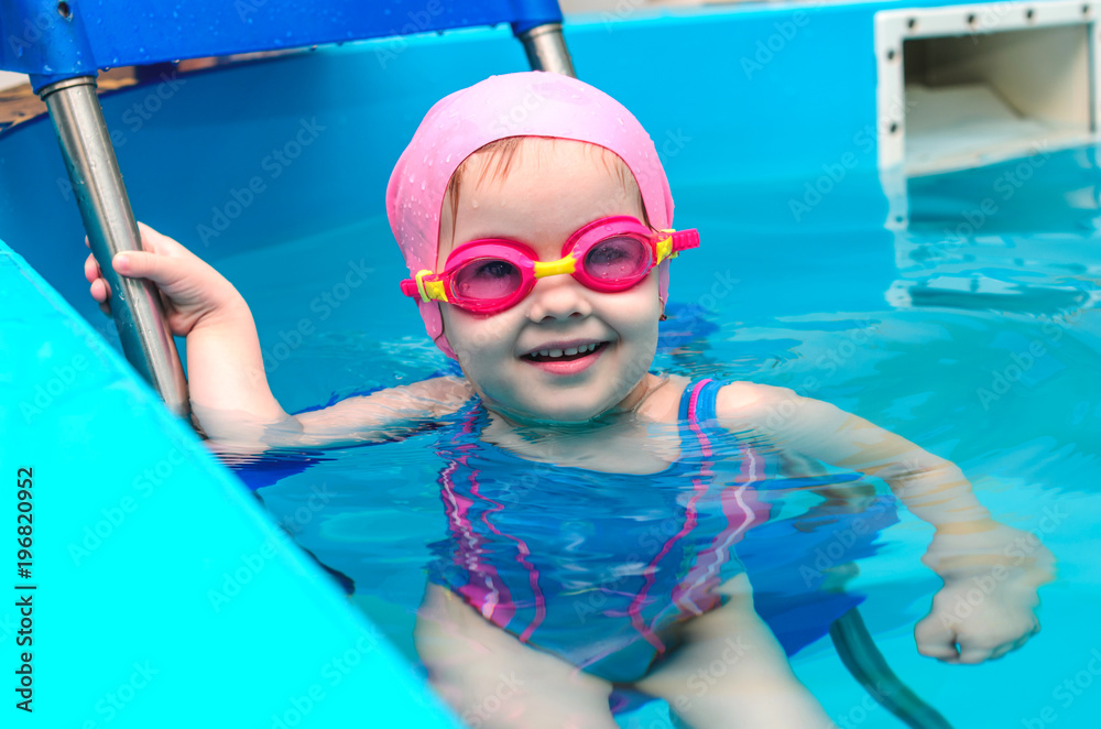 A little girl of European appearance in a pink rubber cap learning to swim in the pool in glasses for swimming