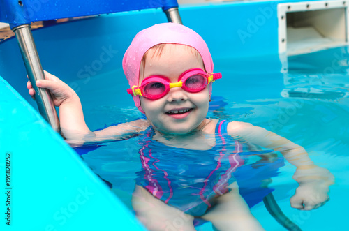 A little girl of European appearance in a pink rubber cap learning to swim in the pool in glasses for swimming