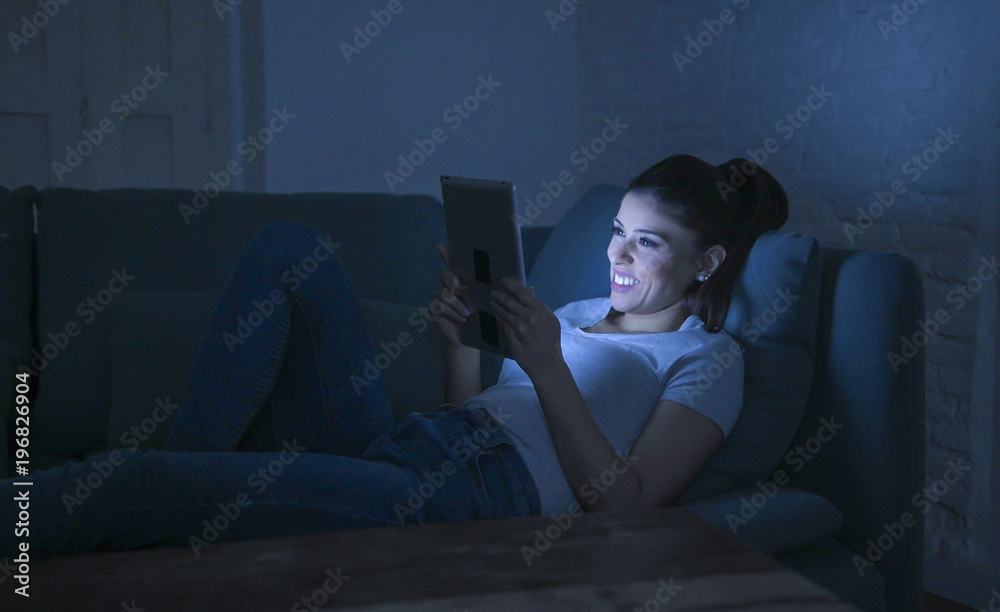Naklejka premium young beautiful happy and relaxed latin woman 30s lying on home couch late night using digital device laptop tablet pad watching internet movie smiling
