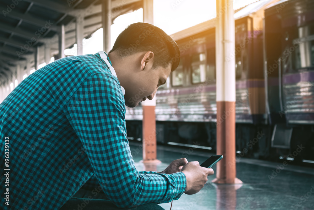 Rear view of young asian hipster man holding mobile phone using app song with linten music at train station. Rear view of young asian hipster man holding mobile phone using app song with linten music at train station.