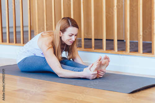 Middle aged blond woman practicing yoga, sitting in Seated forward bend exercise, paschimottanasana pose, working out, wearing sportswear, grey pants, indoor, home interior wooden background,side view photo