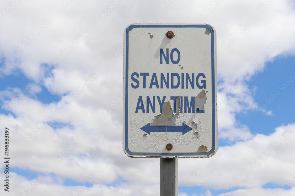 a weathered, peeling blue and white No Standing Any Time sign with double ended arrow in a cloudy sky