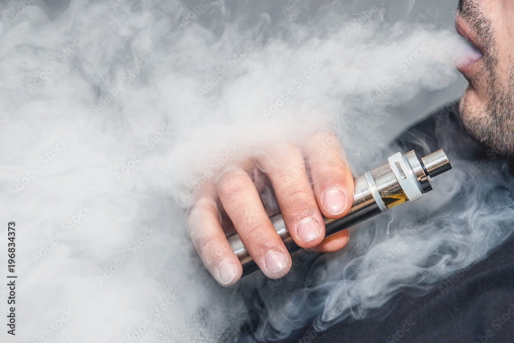 A man smokes an electronic cigarette on a gray background, blowing a stream of smoke. Copy space.