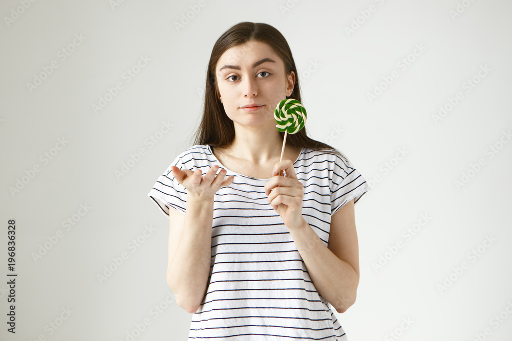 Indoor isolated shot of serious beautiful casually dressed young European woman holding lollipop, gesturing, expressing uncertainty and doubt, being unsure about eating sugar sweet unhealthy food Indoor isolated shot of serious beautiful casually dressed young European woman holding lollipop, gesturing, expressing uncertainty and doubt, being unsure about eating sugar sweet unhealthy food
