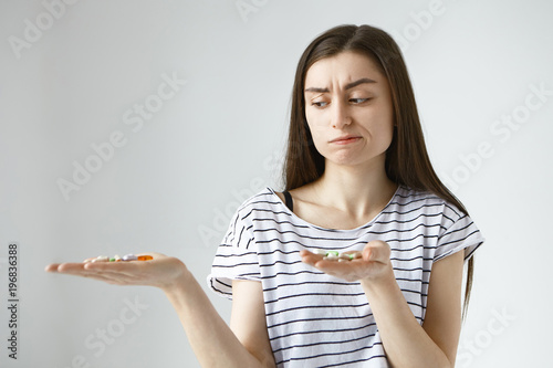 Bilde på lerret Frustrated doubtful young Caucasian female frowning and pursing lips, can't chose which painkillers to take while suffering from cramps